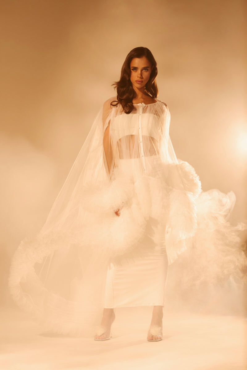 A woman stands in a hazy, softly lit setting wearing the PIPER Soft Tulle Blouse in White with ruffled trim and voluminous details. Her hair is styled in loose waves as she gazes confidently at the camera.