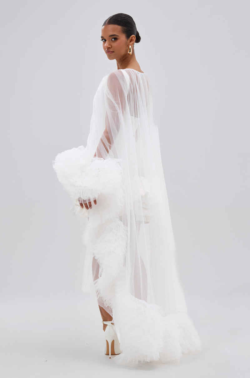 A woman wears the PIPER SOFT TULLE BLOUSE IN WHITE, styled with tulle cuffs and feathered trim. She pairs it with high heels, large gold earrings, and has her hair sleekly pulled back, posing against a plain light background.