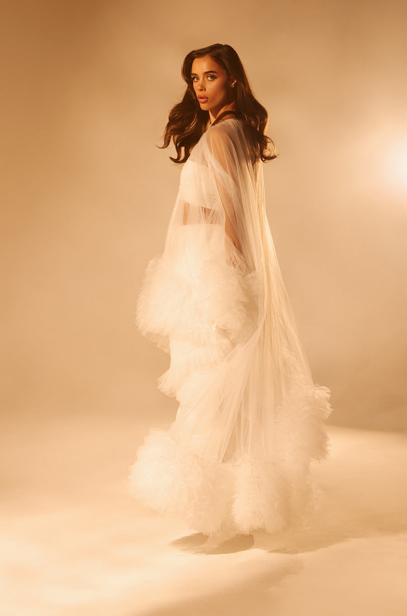 A woman stands in soft, warm light wearing the Piper Soft Tulle Blouse in White, featuring sheer fabric and ruffled trim. Her long hair flows in loose waves as she gazes confidently at the camera.