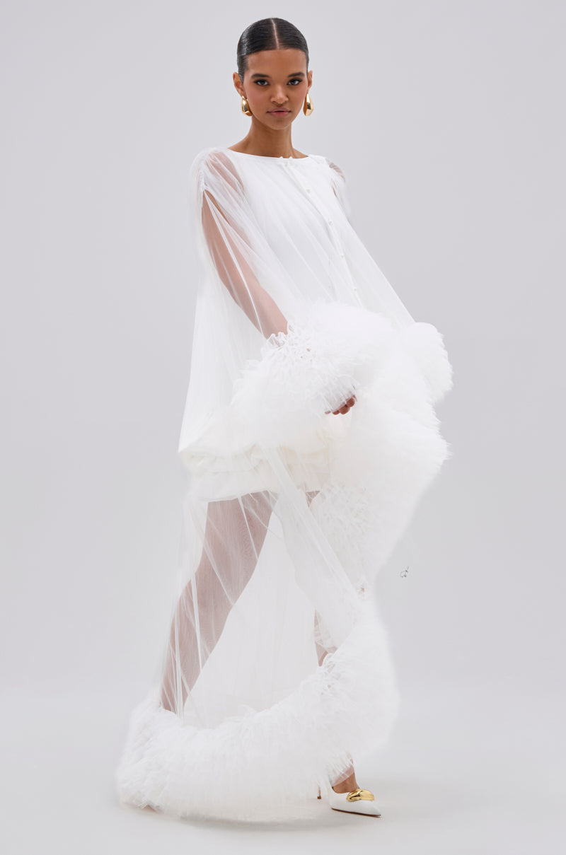 A woman poses against a plain light background in the Piper Soft Tulle Blouse in white, featuring sheer fabric and voluminous ruffled trim at the hem and sleeves, paired with white high heels with gold accents. Her hair is slicked back.