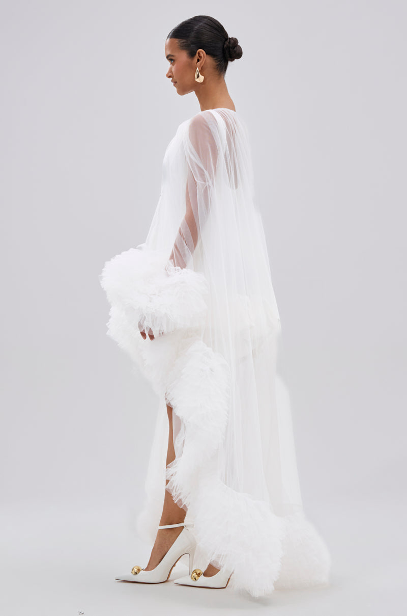 A woman in profile wears the Piper Soft Tulle Blouse in White, featuring ruffled trim at the hem and sleeves. She styles it with a sleek bun, gold earrings, and white heels against a plain, light background.