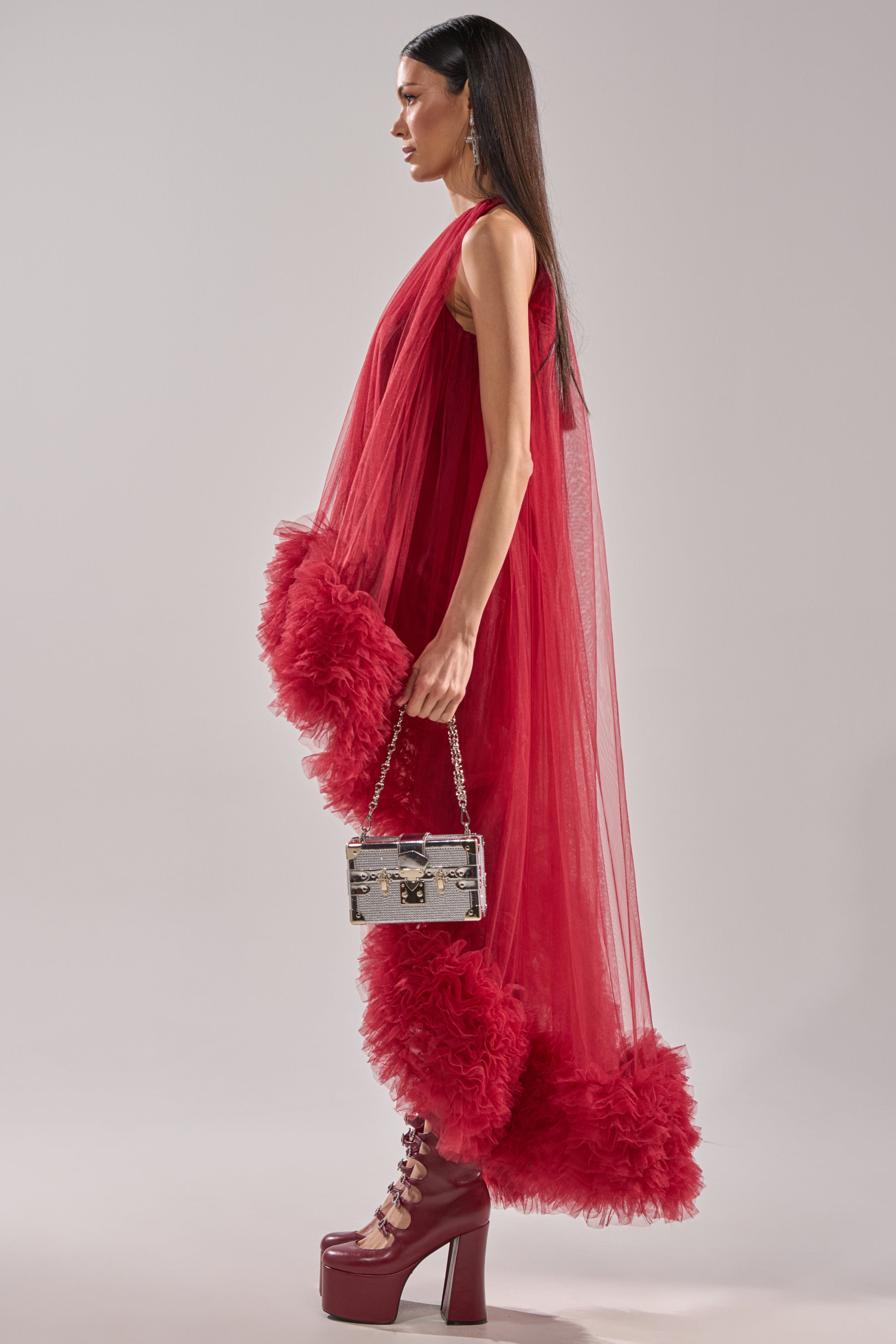 A woman in the ARI TULLE HIGH LOW DRESS IN BURGUNDY with a ruffled hem, matching platform heels, and a small silver chain-strap handbag stands in profile against a plain light background.
