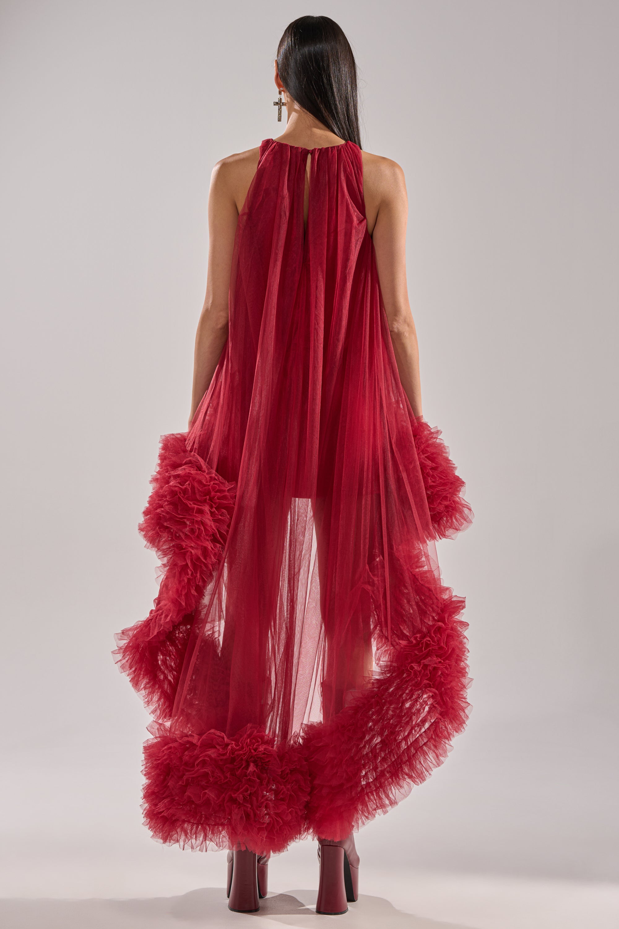 A person with long dark hair faces away, wearing the ARI TULLE HIGH LOW DRESS IN BURGUNDY with dramatic ruffled trim and matching burgundy platform boots, set against a plain light background.