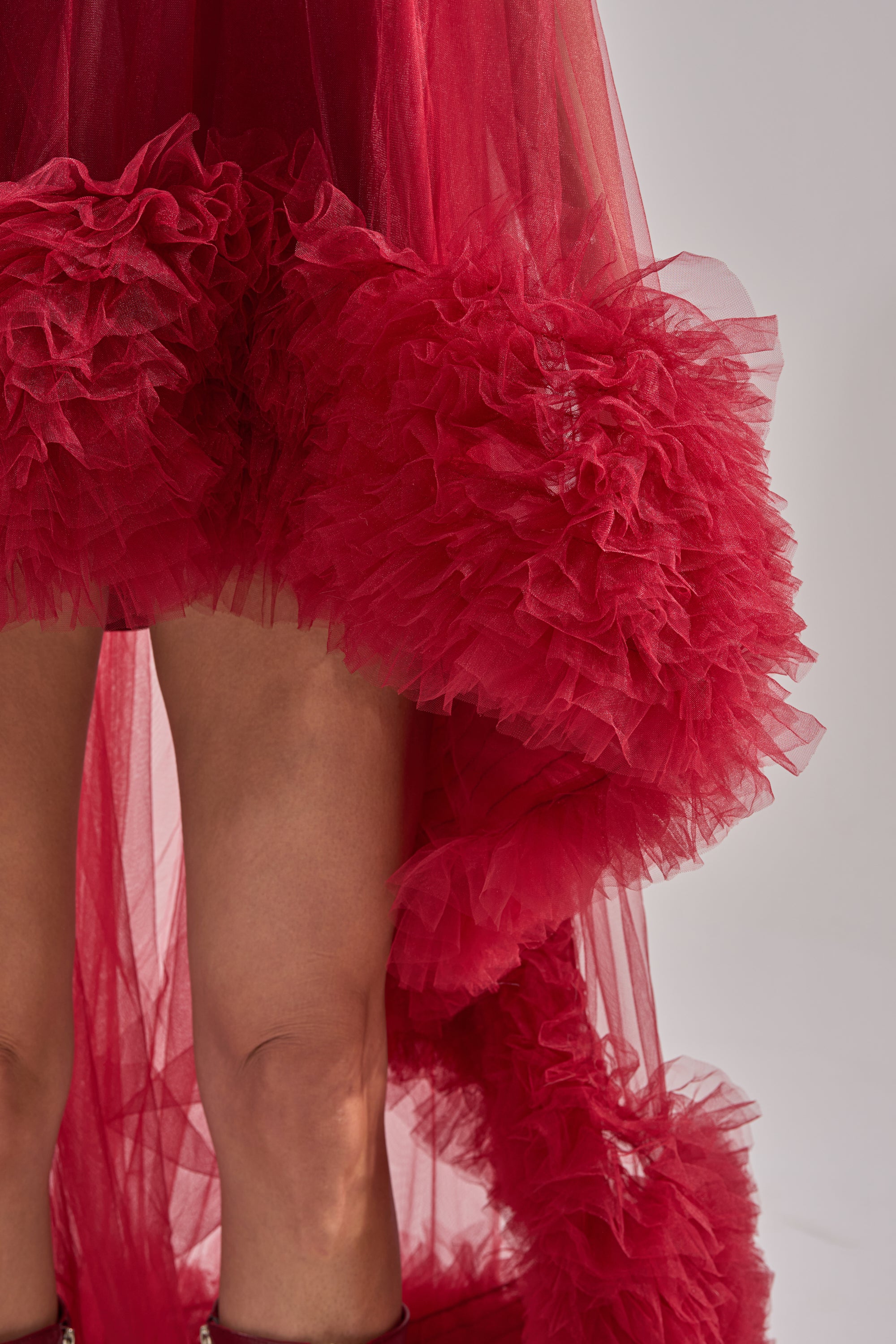 Close-up of a person wearing the ARI TULLE HIGH LOW DRESS IN BURGUNDY, featuring dramatic tulle ruffles around the hem and bare legs. The mini dress stands out against a neutral, softly lit background.