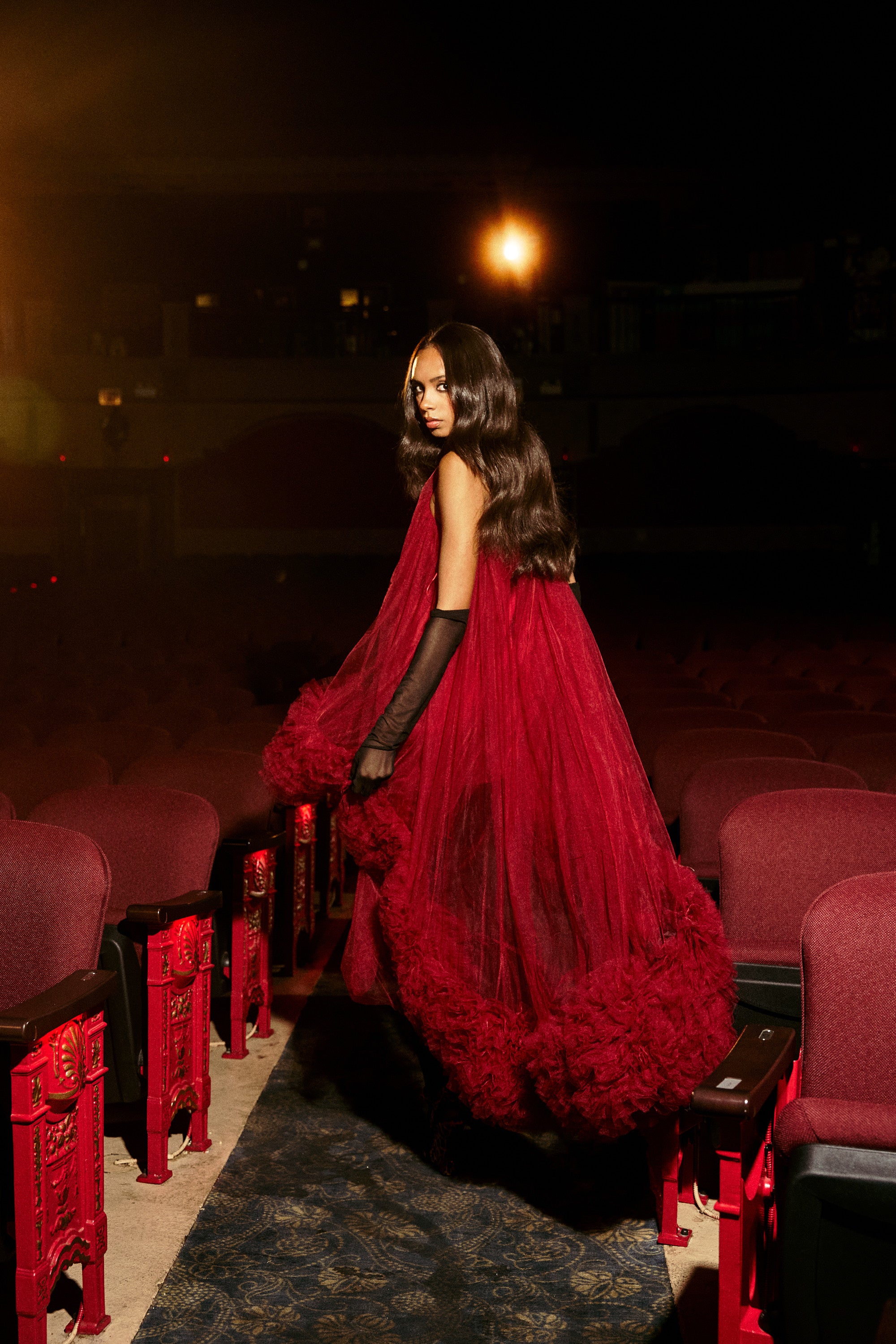 A woman in the ARI TULLE HIGH LOW DRESS IN BURGUNDY stands elegantly between red theater seats, glancing back over her shoulder. Warm lighting emphasizes her flowing hair and the gown's rich texture.