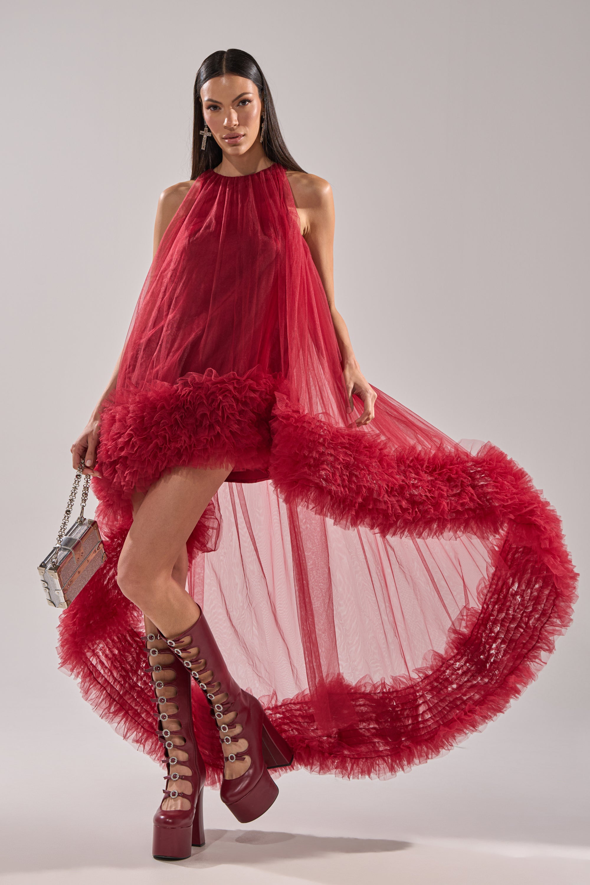 A woman in the ARI TULLE HIGH LOW DRESS IN BURGUNDY with a dramatic sheer hem stands holding a small purse. She wears tall, lace-up red platform boots and has long, straight dark hair.