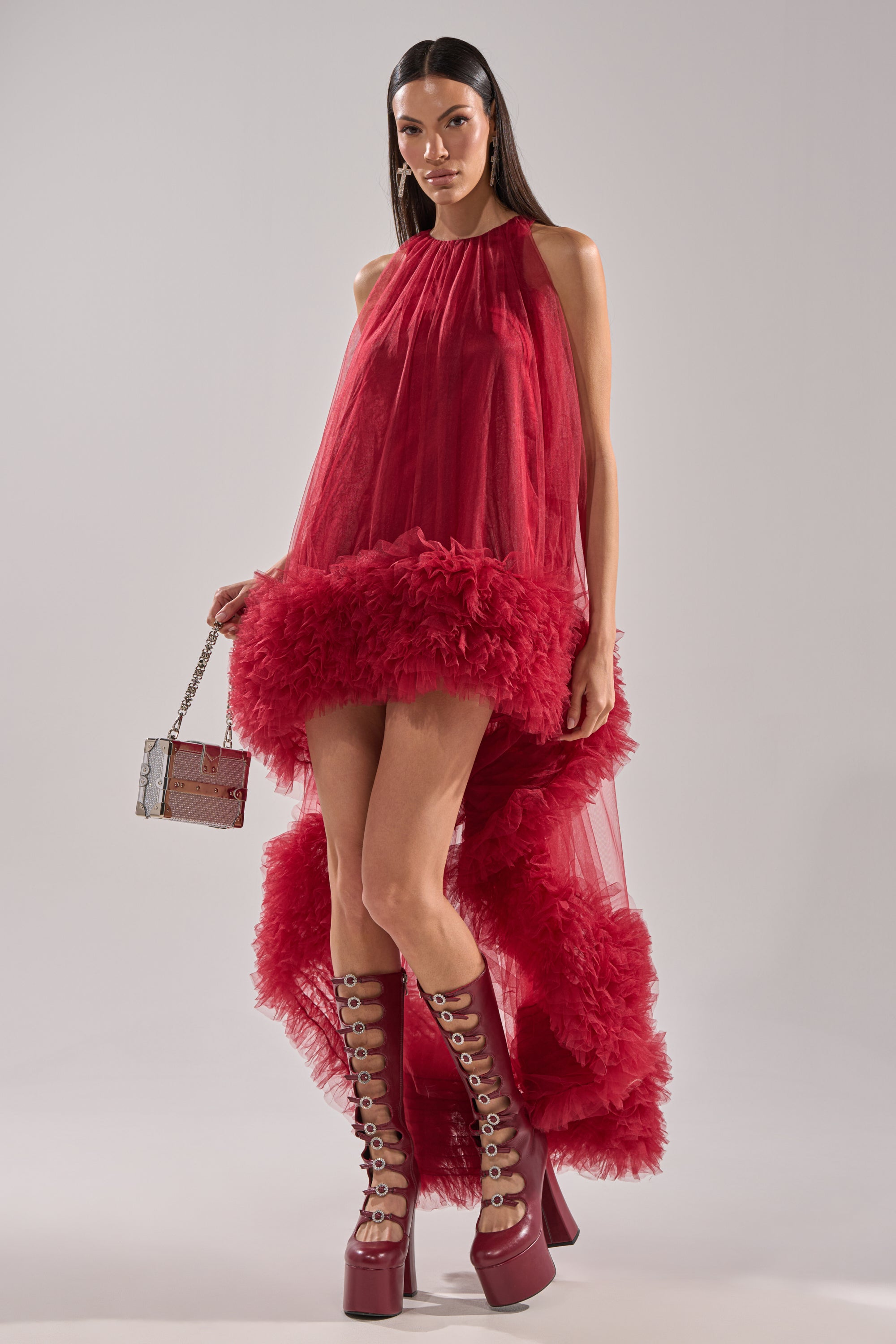 A woman in the ARI TULLE HIGH LOW DRESS IN BURGUNDY with a ruffled hem, platform lace-up boots, and a metallic purse stands against a plain background. Her long dark hair is worn straight.