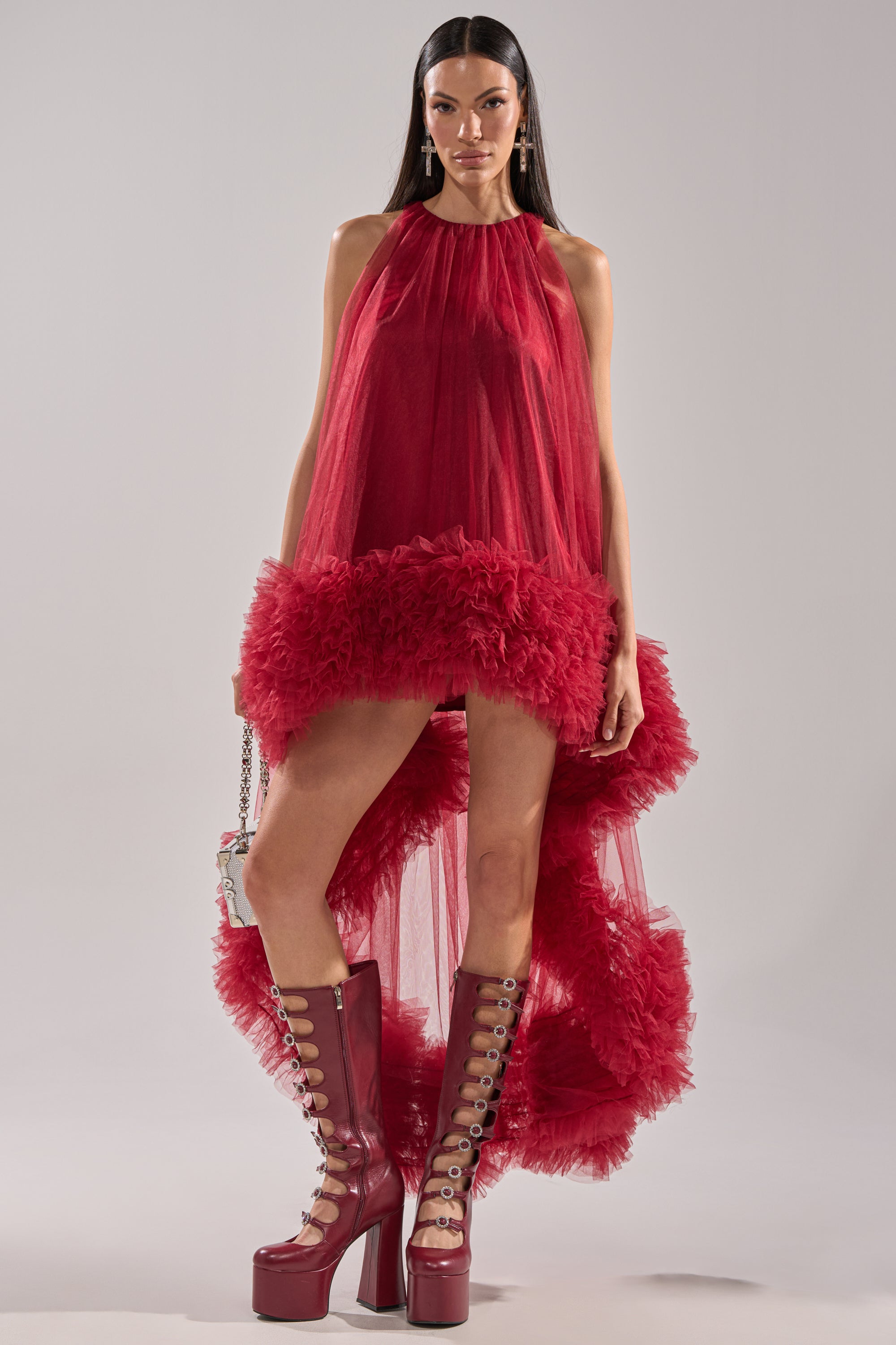 A woman poses confidently against a plain background in the ARI TULLE HIGH LOW DRESS IN BURGUNDY by AKIRA Label, styled with tall lace-up red platform boots and a small handbag.