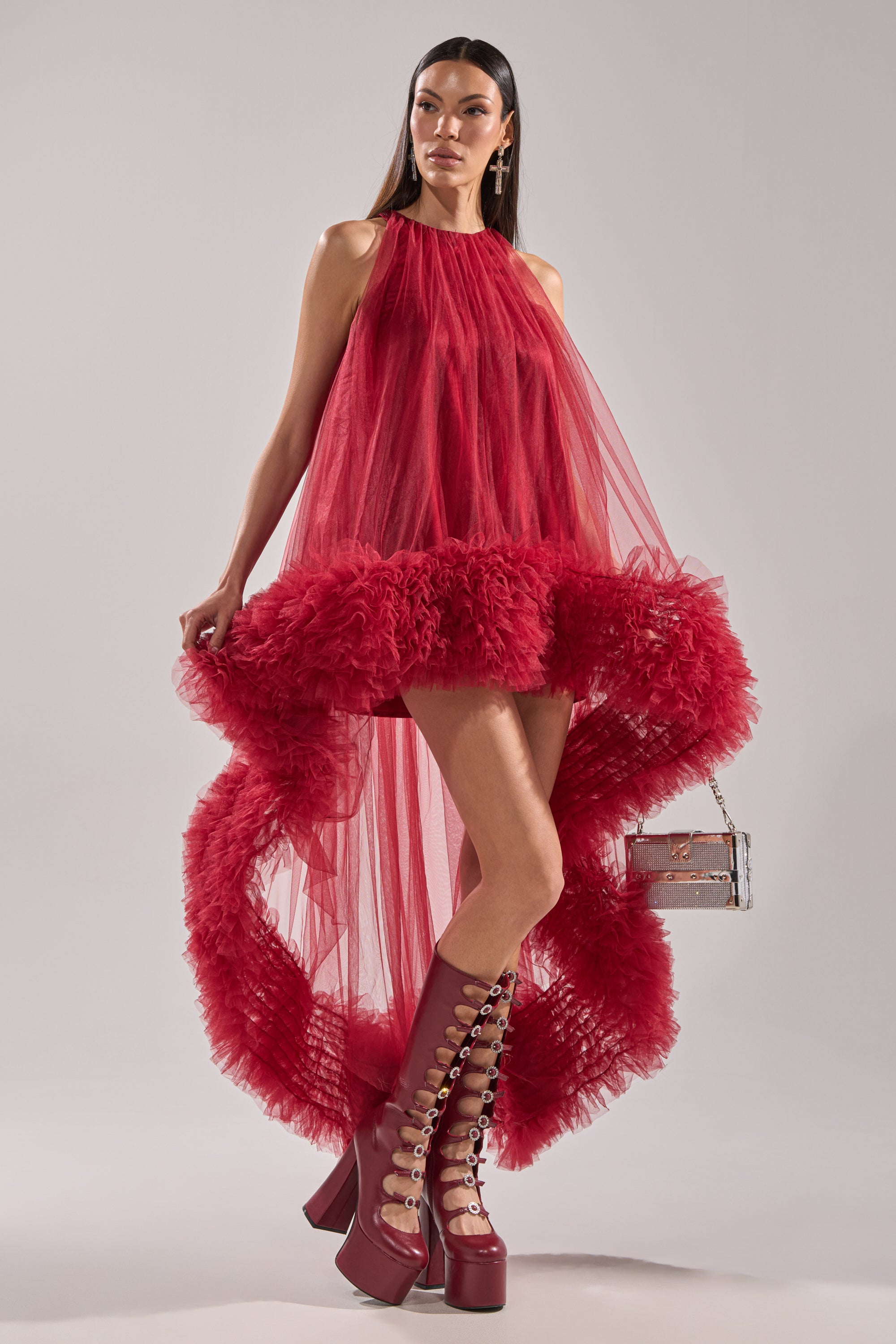 A woman in the ARI TULLE HIGH LOW DRESS IN BURGUNDY with a dramatic ruffled hem and tall lace-up platform boots holds a small purse, standing confidently against a plain background.