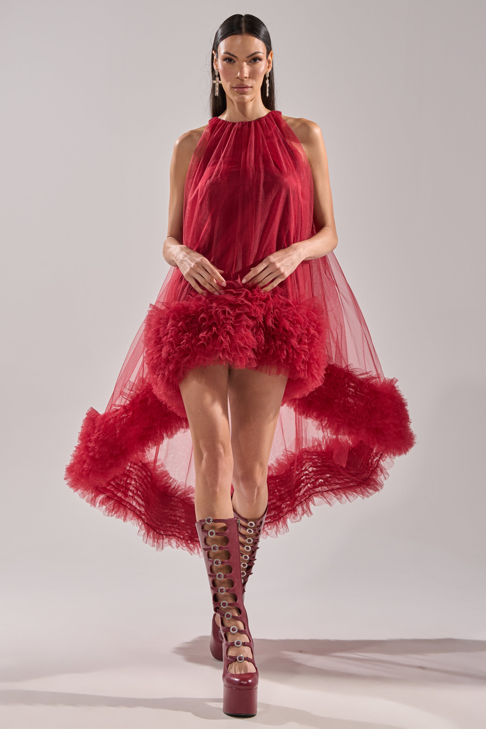 A woman wears the ARI TULLE HIGH LOW DRESS IN BURGUNDY with a sheer top and feathered skirt, pairing it with red lace-up platform boots and bold earrings for an edgy party vibe.