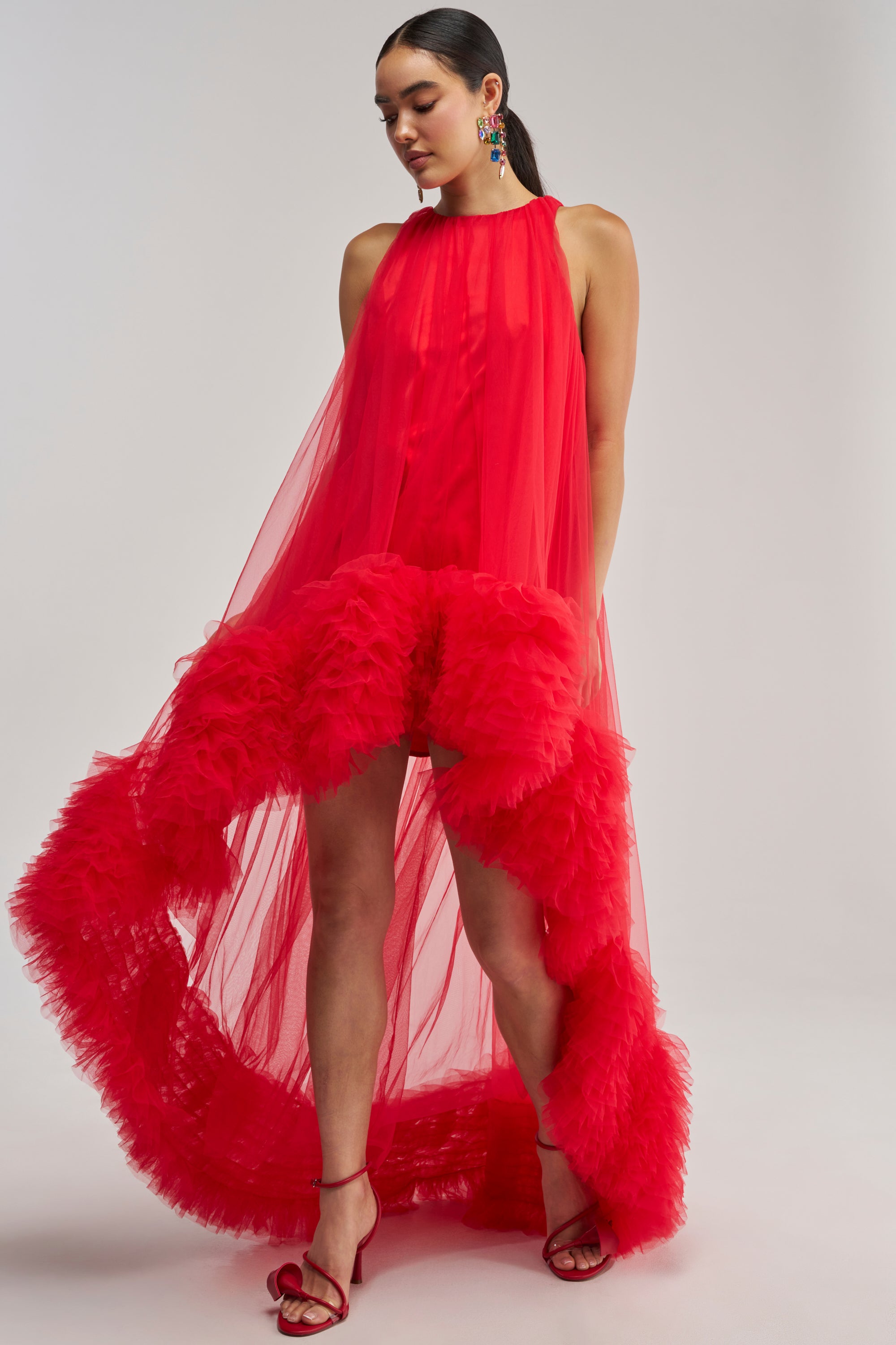 Wearing the ARI TULLE RUFFLE HIGH LOW MINI DRESS IN RED—a vibrant high-low mini featuring a sheer tulle top layer and dramatic ruffled hem—she completes her special event look with red strappy heels and colorful earrings, posing confidently against a plain light background.