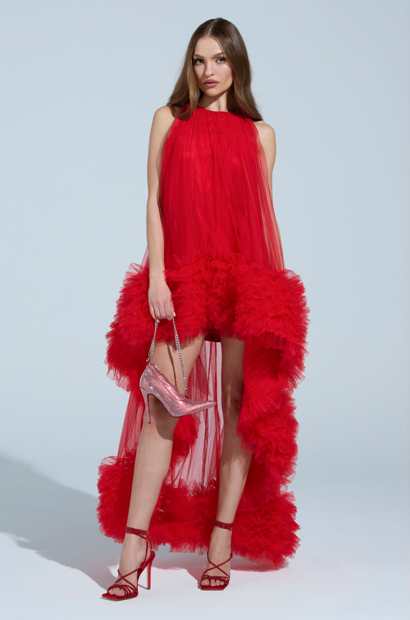 Wearing the ARI tulle ruffle high low mini dress in red, a person holds a sparkling red high heel. They pair it with matching red strappy heels, standing against a plain background while their long, straight hair completes the elegant look.