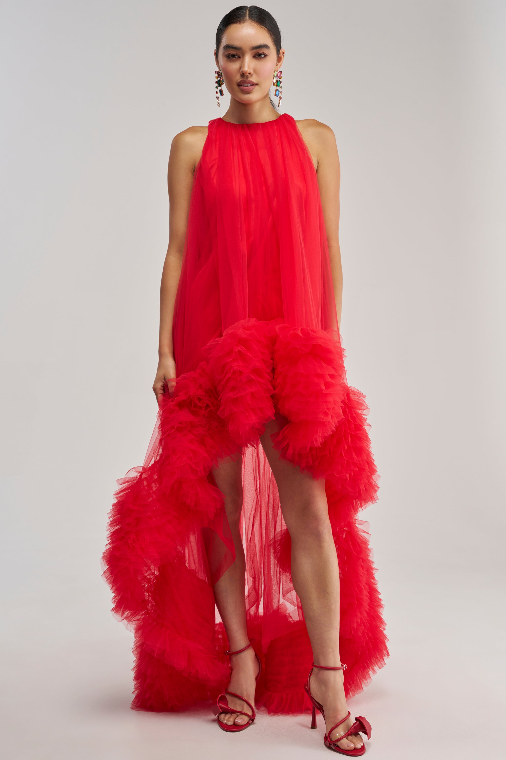 A woman stands against a plain background wearing the ARI TULLE RUFFLE HIGH LOW MINI DRESS IN RED, a sleeveless, sheer mini dress featuring dramatic layered ruffle details and a high-low hem. Styled with red heels and long, colorful earrings, her dark hair is pulled back—making this eye-catching piece the perfect choice for any special event or occasion.