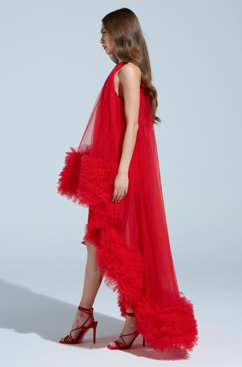 A woman in profile wears the ARI Tulle Ruffle High Low Mini Dress in red, featuring a tulle ruffle and a dramatic train. Her long, wavy hair and red high-heeled sandals complete the undeniably red-carpet-ready look against a light gray backdrop.