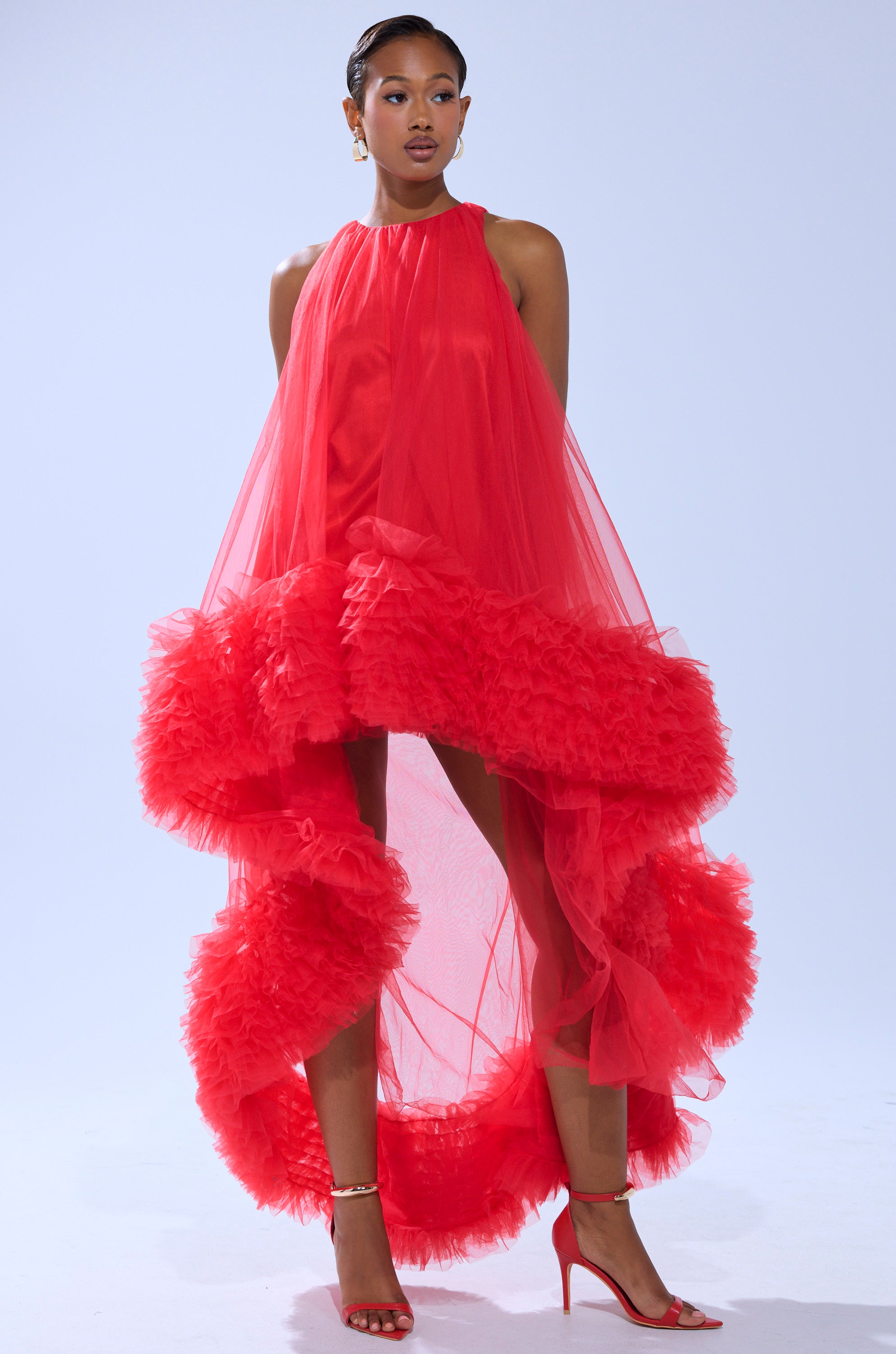 A woman models the ARI TULLE RUFFLE HIGH LOW MINI DRESS IN RED, featuring dramatic voluminous ruffles and a high-low hem, paired with matching red heels against a light blue background.