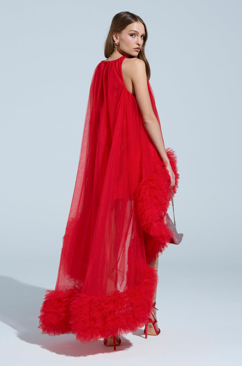 A woman models the ARI TULLE RUFFLE HIGH LOW MINI DRESS IN RED, featuring a sheer tulle overlay and ruffled hem. Against a light blue backdrop, she holds a small silver clutch. Her long hair cascades down, paired with striking red high heels for an elegant look.
