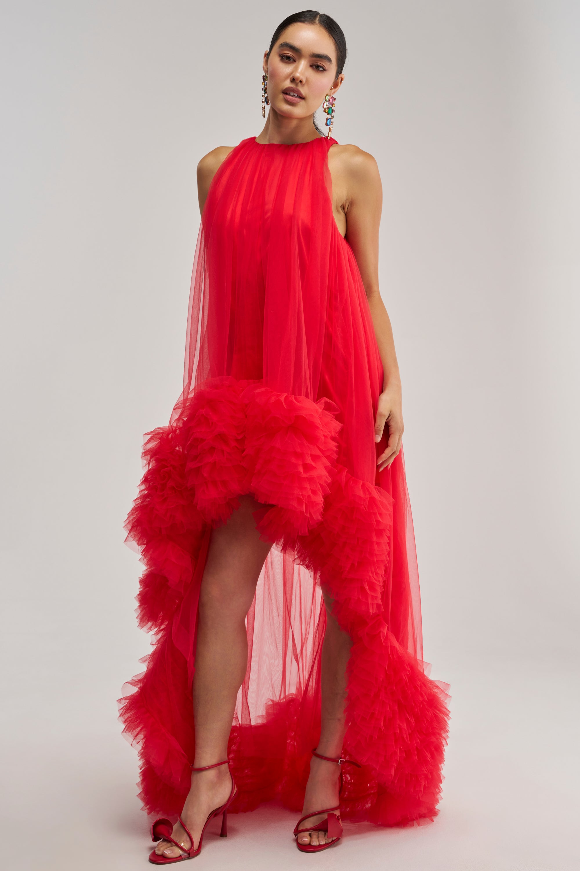 A woman poses in the ARI TULLE RUFFLE HIGH LOW MINI DRESS IN RED, a sleeveless, sheer mini dress with a high neckline and dramatic ruffled high-low hem, styled with red heels and colorful earrings against a plain background.