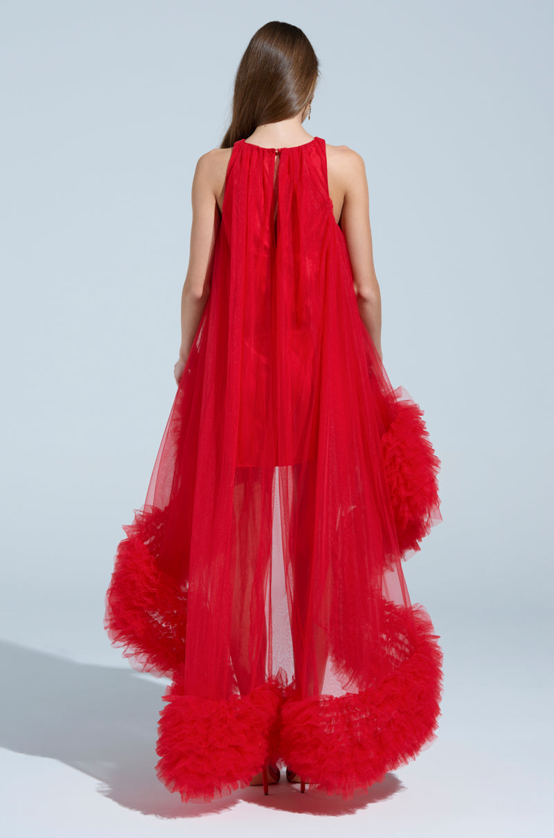 Wearing the ARI TULLE RUFFLE HIGH LOW MINI DRESS IN RED, a person stands with their back turned. The flowing sheer fabric and voluminous tulle ruffles at the hem are contrasted against a light backdrop, making them red-carpet ready.