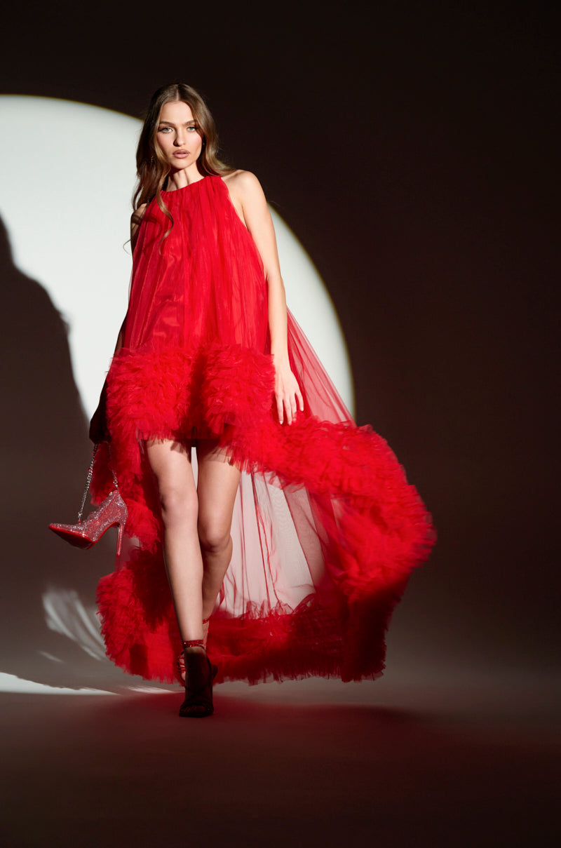 In the ARI Tulle Ruffle High Low Mini Dress in Red, a woman confidently walks, holding high-heeled shoes. She looks red-carpet ready as she stands silhouetted against a circular light.
