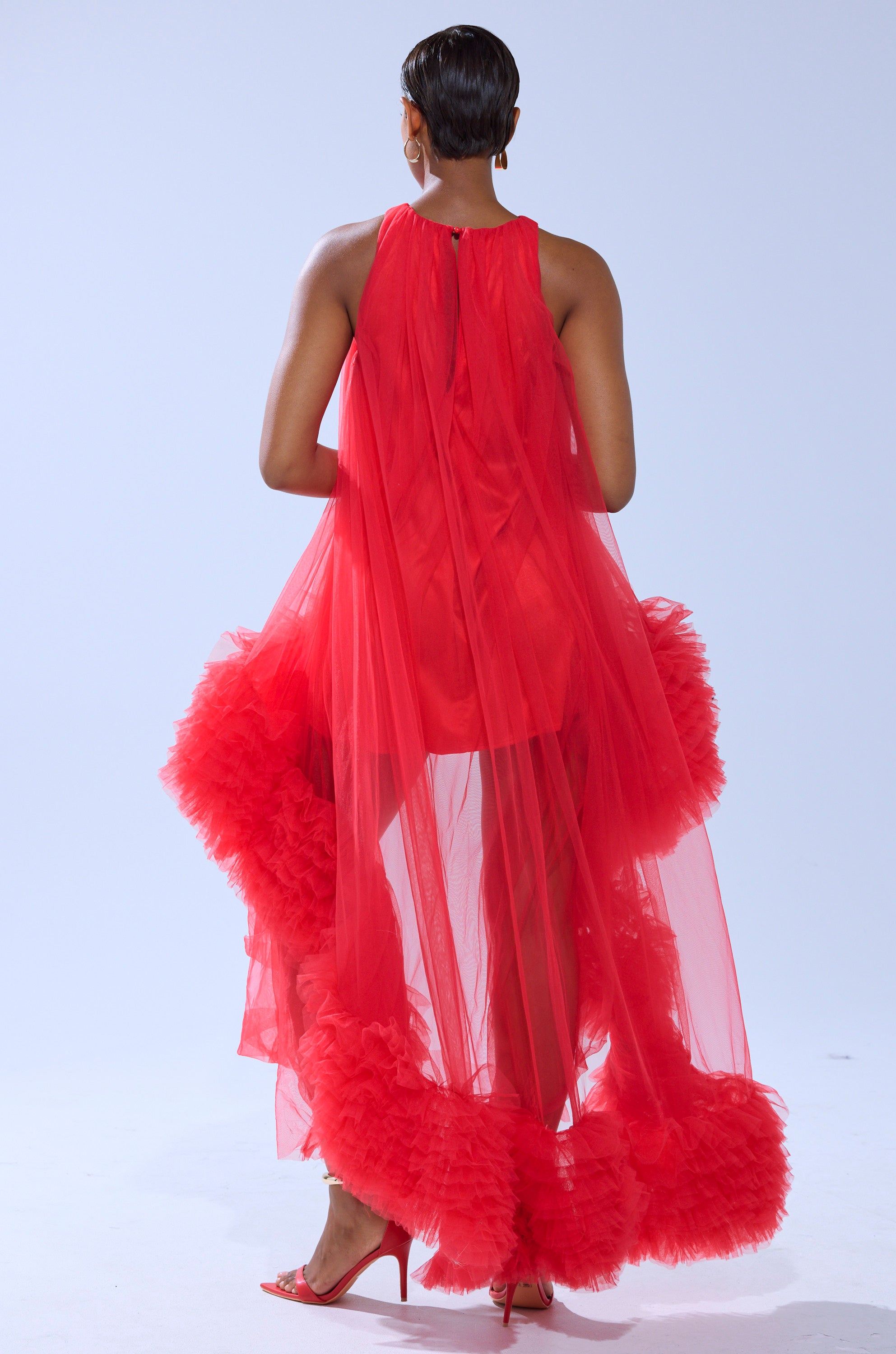 Someone stands facing away in the ARI TULLE RUFFLE HIGH LOW MINI DRESS IN RED, the sheer ruffled hem and bold color trailing elegantly behind them, set against a plain background.
