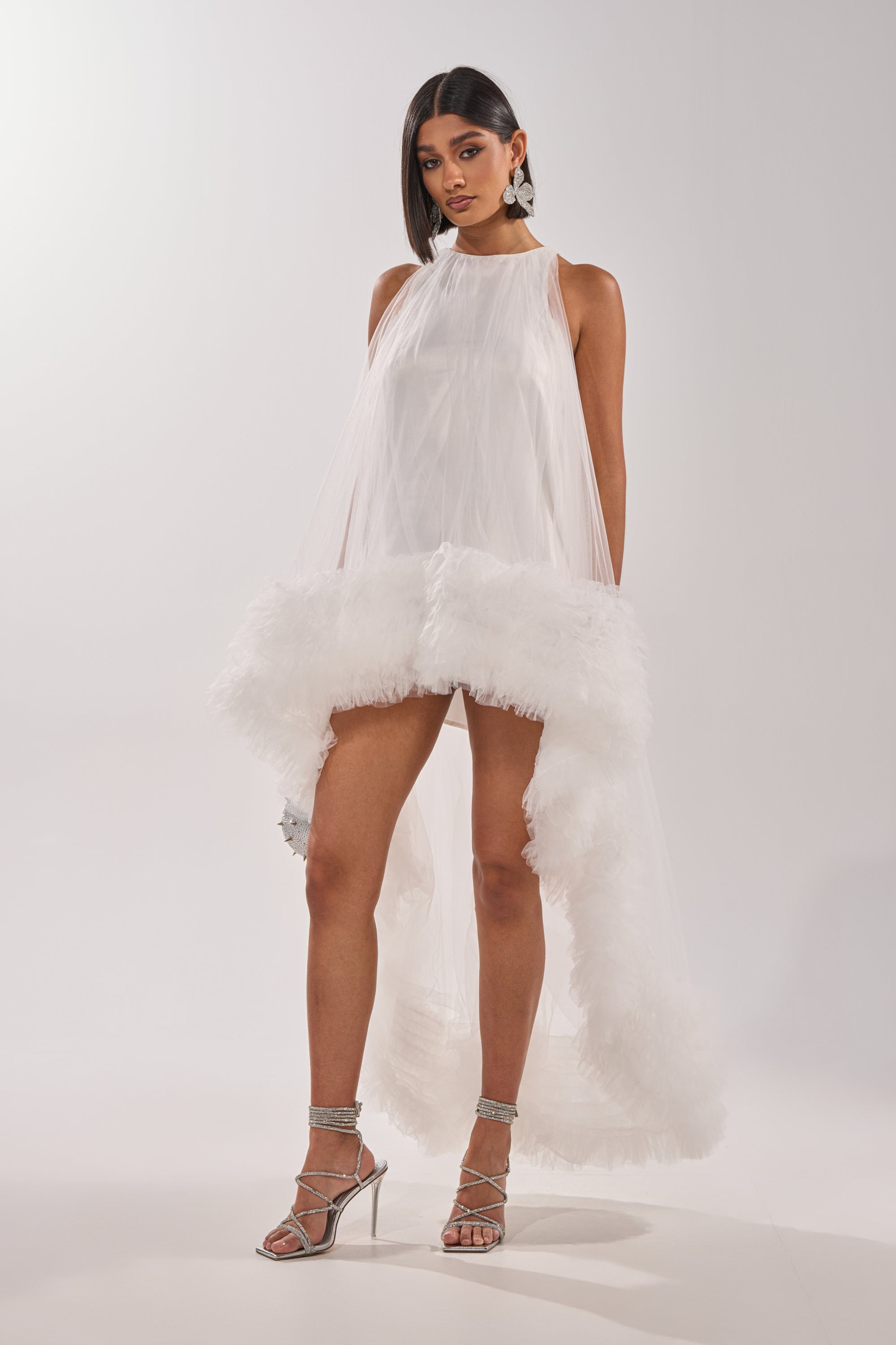A woman wears the ARI TULLE RUFFLE HIGH LOW MINI DRESS IN WHITE—sleeveless with a sheer overlay and ruffled hem—paired with silver high-heeled sandals and statement earrings, standing against a plain white background.