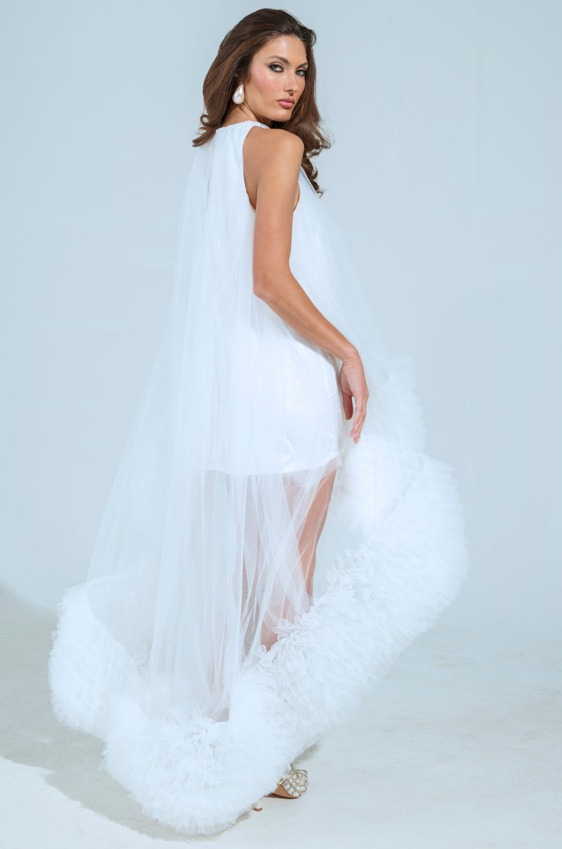 A woman poses confidently in a studio wearing the AKIRA Label's ARI TULLE RUFFLE HIGH LOW MINI DRESS IN WHITE, adorned with a sheer cape and feathered hem. She pairs it with large earrings and strappy heels, glancing over her shoulder against a plain background.