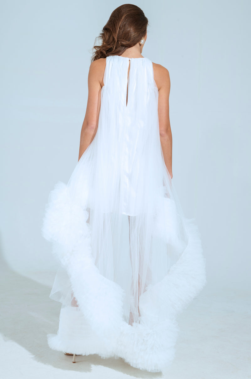 A person in a flowing ARI TULLE RUFFLE HIGH LOW MINI DRESS IN WHITE stands with their back to the camera, set against a plain background. This piece is long and elegant, featuring textured details at the hem for added sophistication.
