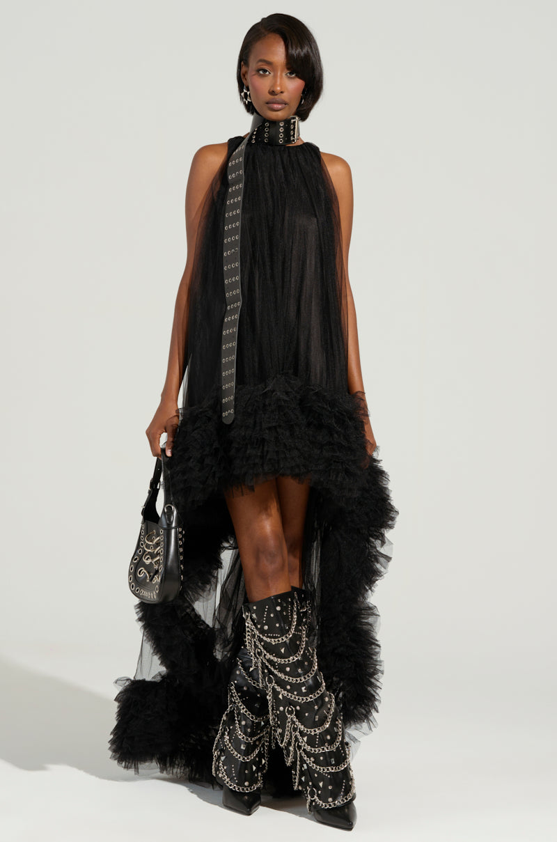 A person confidently stands wearing the ARI Tulle Ruffle High Low Dress in black, featuring a high collar and tulle ruffle hem. They have a matching handbag and knee-high boots with intricate chain detailing. The plain background accentuates their distinctive fashion.