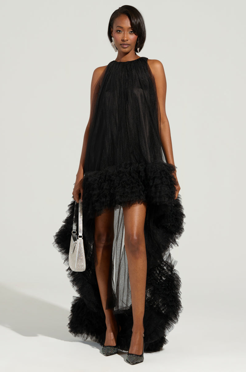 A person stands confidently in the ARI TULLE RUFFLE HIGH LOW DRESS IN BLACK, a sleeveless ensemble featuring elegant tulle ruffles. They hold a silver handbag and wear sleek black pointed heels, perfectly complementing the stylish outfit against a plain background.