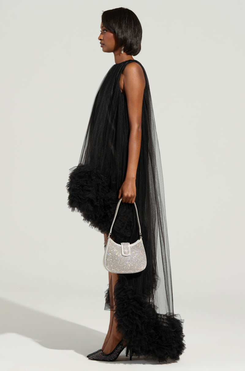 A woman in the ARI TULLE RUFFLE HIGH LOW DRESS IN BLACK, elegantly styled with voluminous tulle ruffle details, poses in profile. She's accessorizing with a sparkling white handbag and polka-dotted black heels against a simple, soft gray backdrop.