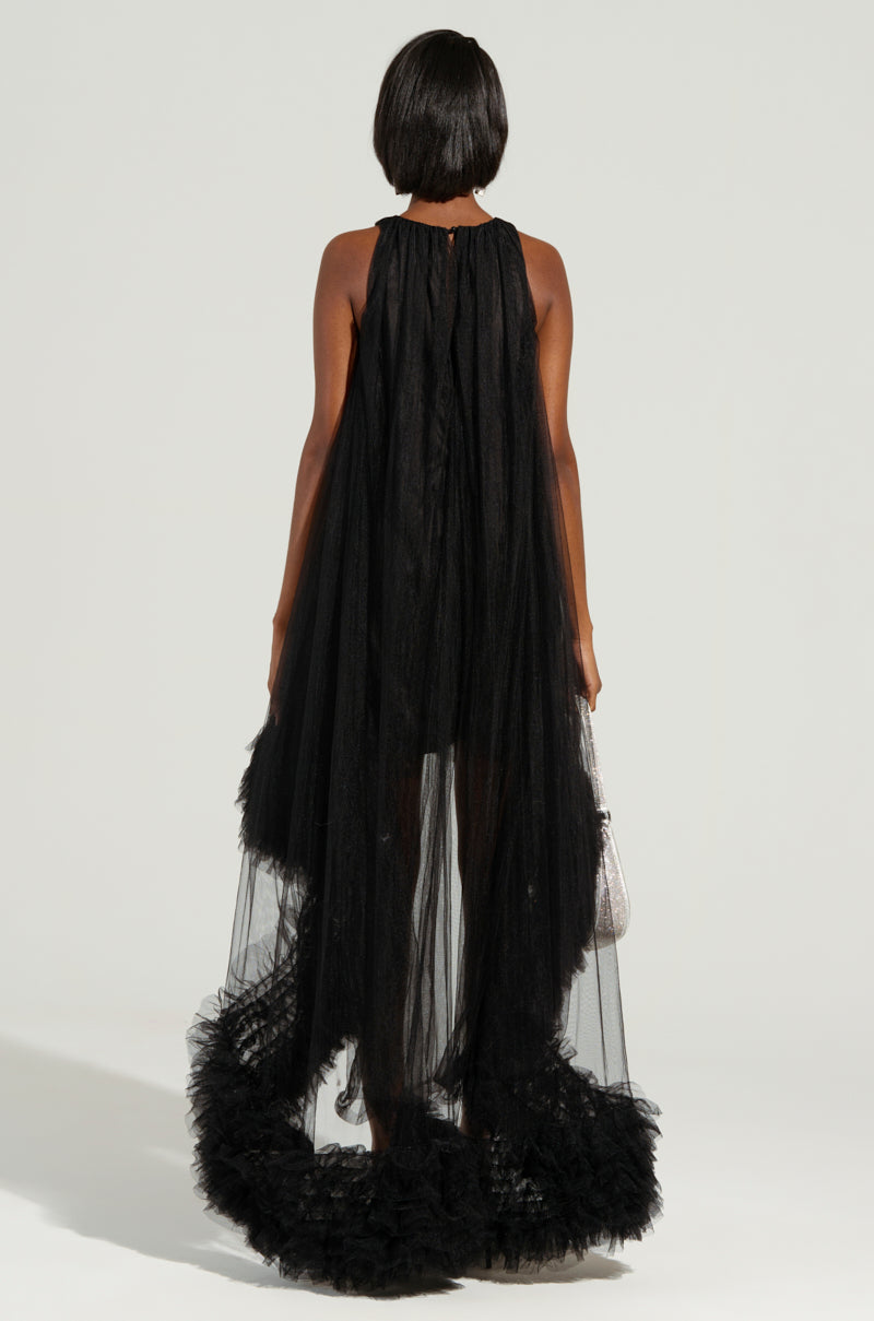 The individual stands with their back turned, draped in an ARI Tulle Ruffle High Low Dress in Black, characterized by its long, flowing silhouette and tulle ruffles lining the edges. They clutch a small handbag and sport short black hair, against a neutral gray backdrop that enhances the scene's timeless elegance.