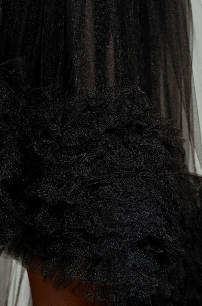 A close-up of the ARI TULLE RUFFLE HIGH LOW DRESS IN BLACK showcases the layered tulle ruffles at the bottom, highlighting its texture and intricate details. The image beautifully captures the delicate nature of the material.