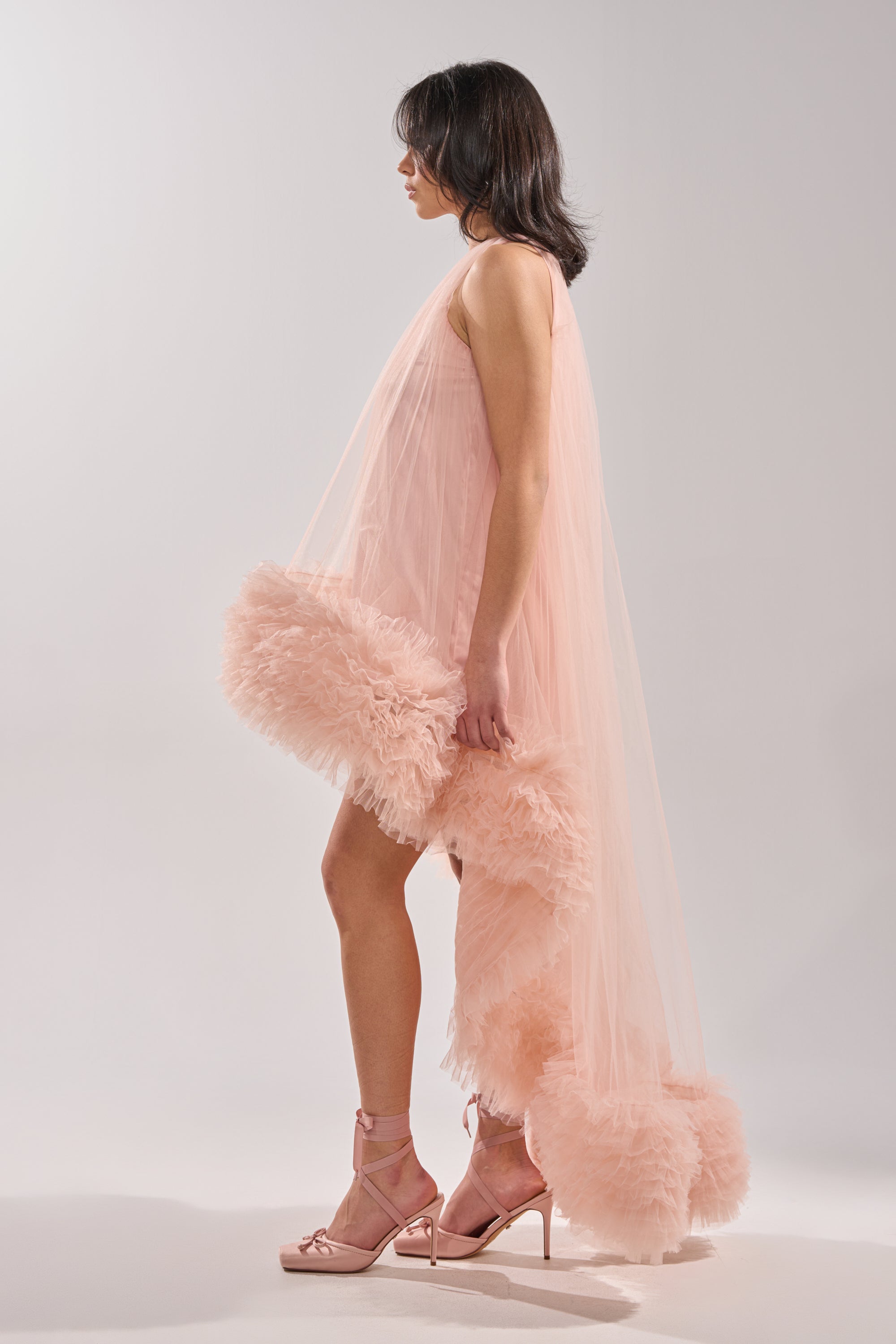A woman stands in profile wearing the ARI TULLE RUFFLE HIGH LOW MINI DRESS IN BLUSH, featuring sleeveless design and voluminous ruffles at the hem, paired with pink high-heeled sandals against a plain light background.