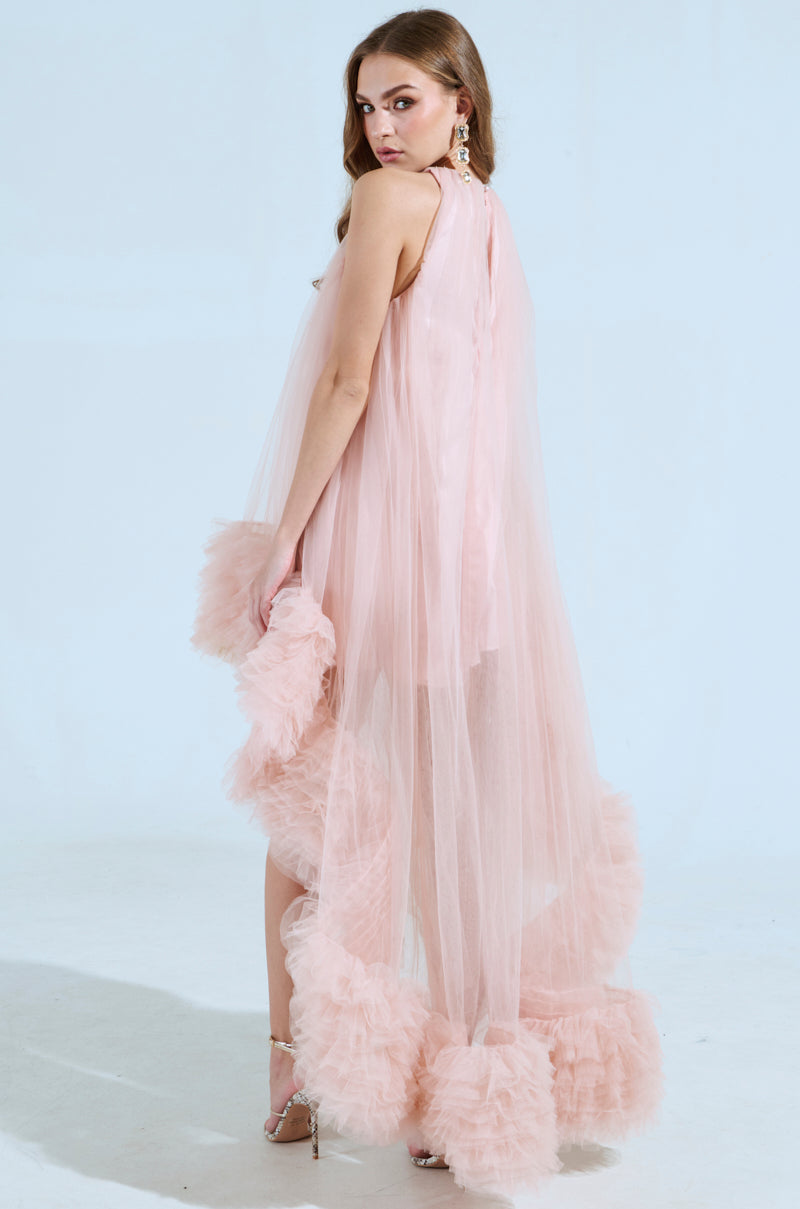 A woman stands elegantly in the ARI TULLE RUFFLE HIGH LOW MINI DRESS IN BLUSH, featuring sheer pink tulle and ruffled edges, with a satin lining. She pairs it with strappy heels and dangling earrings, her long hair cascading down her back as she poses against a plain background.