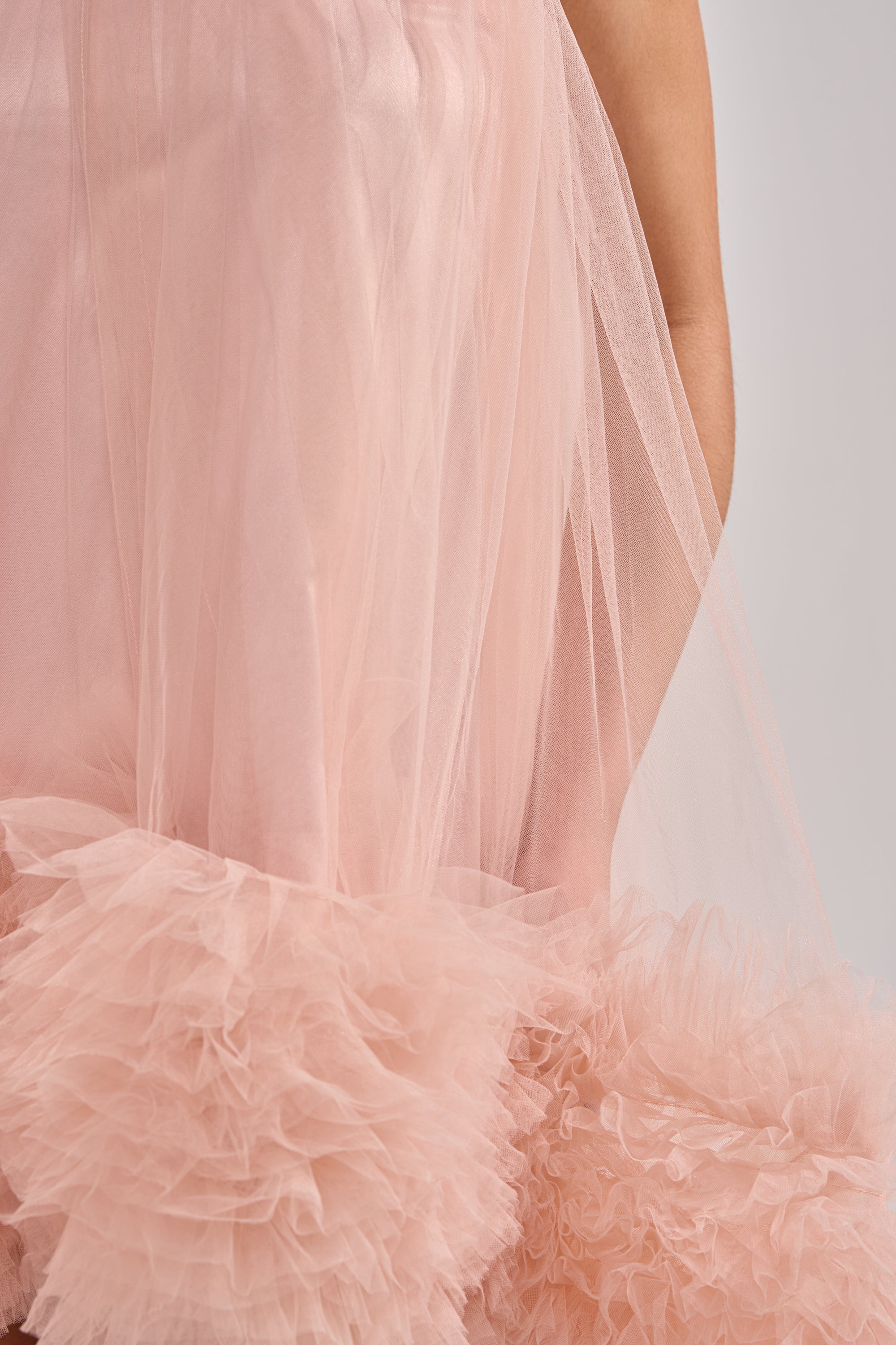 A close-up of the ARI TULLE RUFFLE HIGH LOW MINI DRESS IN BLUSH, featuring fluffy, layered tulle ruffles at the hem. An arm is partially visible against a plain, light background.