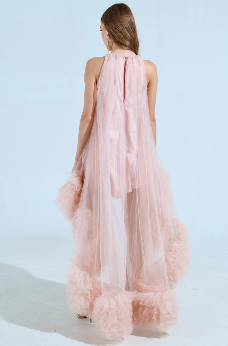 A woman faces away from the camera in an ARI Tulle Ruffle High Low Mini Dress in Blush, showcasing its flowing design with a sheer pink overlay and satin lining. The dress exudes elegance with its ethereal look and ruffled hem, while her long hair cascades gracefully.