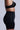 A person is shown from the side wearing a black bra and Titsy Tummy Tuck Shorts in Black, featuring seamless hems that enhance the sleek design, all set against a simple light gray background.