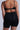 Wearing Titsy Tummy Tuck Shorts in black with seamless hems and a high-rise fit, the person complements a black bra. Their arms feature small tattoos as they pose against a plain gray background.