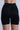 A person is shown from behind wearing TITSY TUMMY TUCK SHORTS in black, featuring seamless hems. The high-waisted shorts offer shaping and compression, extend to mid-thigh, and provide a smooth fit. A plain light background highlights the sleek silhouette.