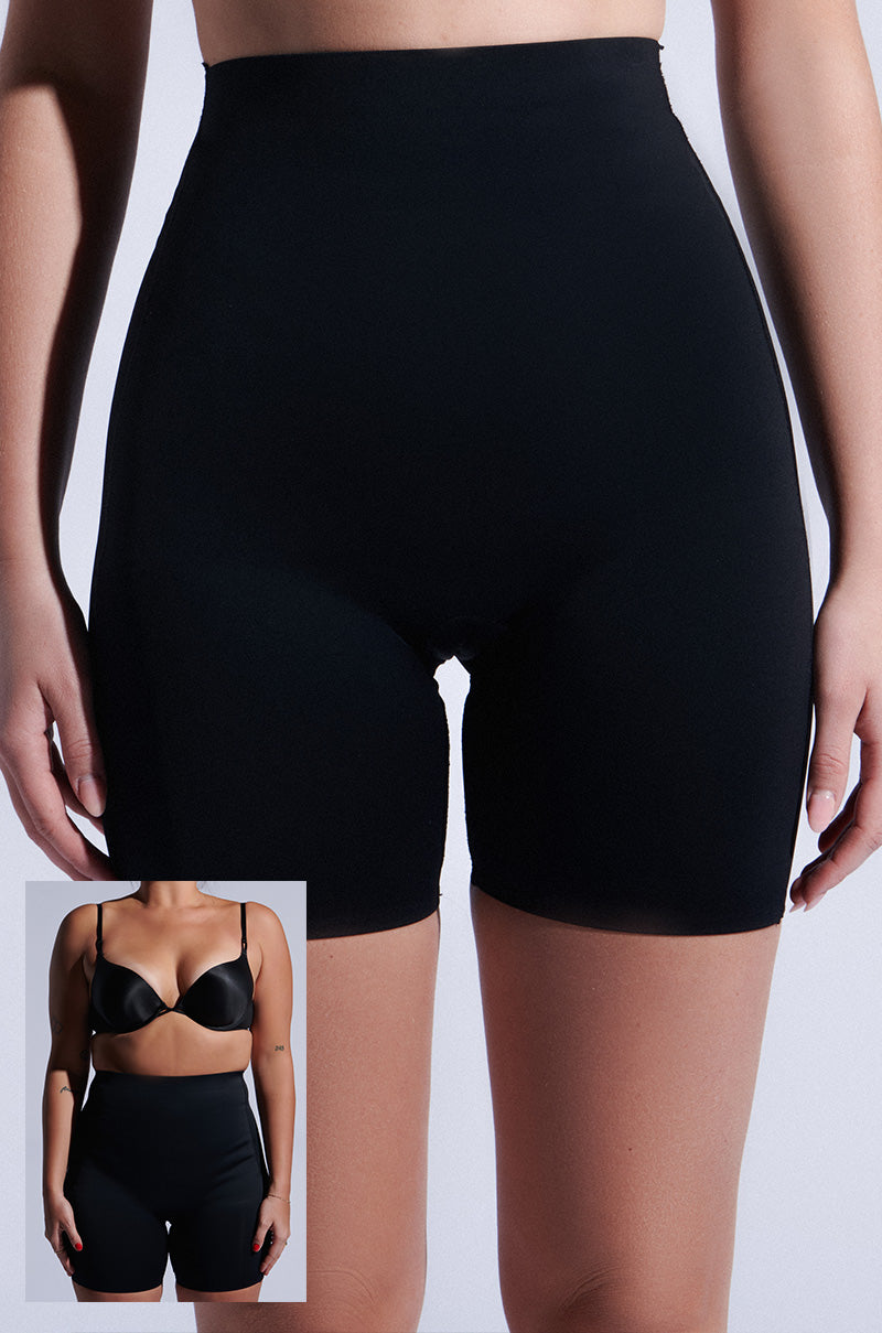 A person wears TITSY TUMMY TUCK SHORTS IN BLACK. The black high-waisted shorts have seamless hems and short legs, paired with a black bra in an inset image. Its high-rise fit provides sleek support against a plain white background.