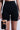 A person wears TITSY TUMMY TUCK SHORTS IN BLACK. The black high-waisted shorts have seamless hems and short legs, paired with a black bra in an inset image. Its high-rise fit provides sleek support against a plain white background.