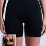 A person wears TITSY TUMMY TUCK SHORTS IN BLACK. The black high-waisted shorts have seamless hems and short legs, paired with a black bra in an inset image. Its high-rise fit provides sleek support against a plain white background.