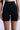 Someone wears TITSY TUMMY TUCK SHORTS IN BLACK, high-waisted with seamless hems reaching mid-thigh. The sleek, form-fitting shapewear sits snugly near the waist against a plain, light-colored background, with the person's arms resting visibly by their sides.