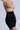 Wearing Titsy Tummy Tuck Shorts in black, someone stands against a light gray background. Their nude bra complements the seamless high-waisted shorts. Their left hand is on their hip while their right arm partially reaches upward.