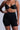 A person wearing TITSY TUMMY TUCK SHORTS IN BLACK and a black bra, with red nail polish, multiple rings, and a bracelet. The background is plain gray.