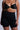A person is wearing TITSY TUMMY TUCK SHORTS IN BLACK with seamless hems and a matching black bra. They have red nail polish and an upper arm tattoo. The background is a simple, neutral color.