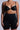 A person in TITSY TUMMY TUCK SHORTS IN BLACK and a black bra stands facing forward. Their tattooed arms rest by their sides against a neutral gradient background.