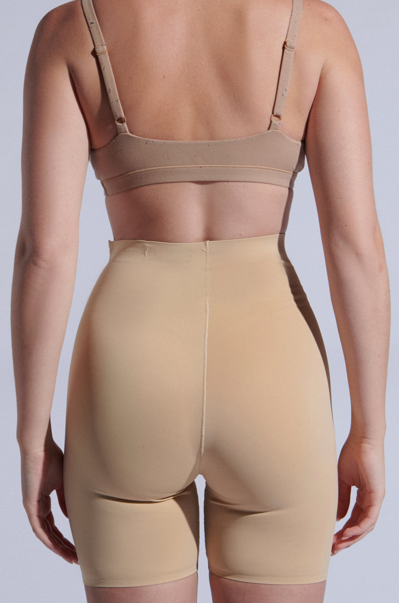 Back view of a person wearing TITSY TUMMY TUCK SHORTS IN TAN and a beige bra, standing against a neutral background. 10