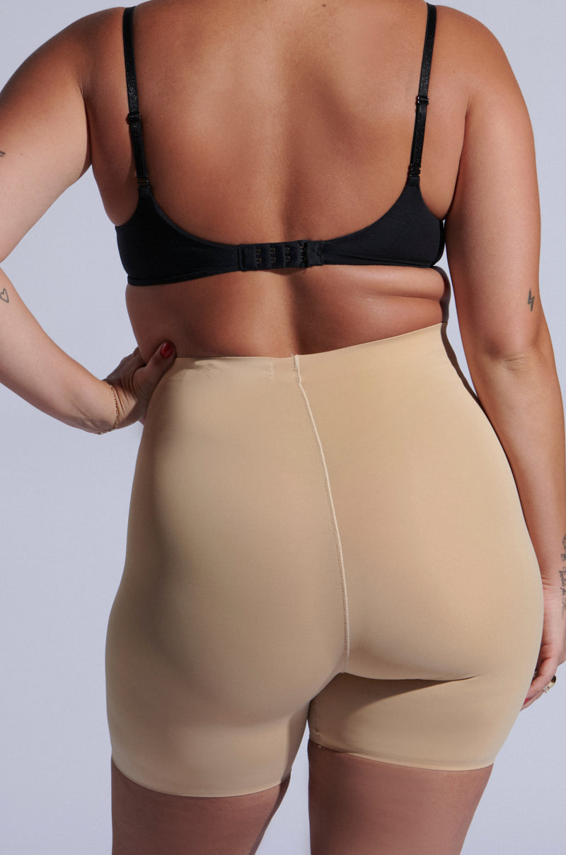 A person is shown from the back wearing TITSY TUMMY TUCK SHORTS IN TAN and a black bra. The image highlights the smooth fabric, high-rise fit, and seamless hems of the shorts against a plain, neutral background. 11