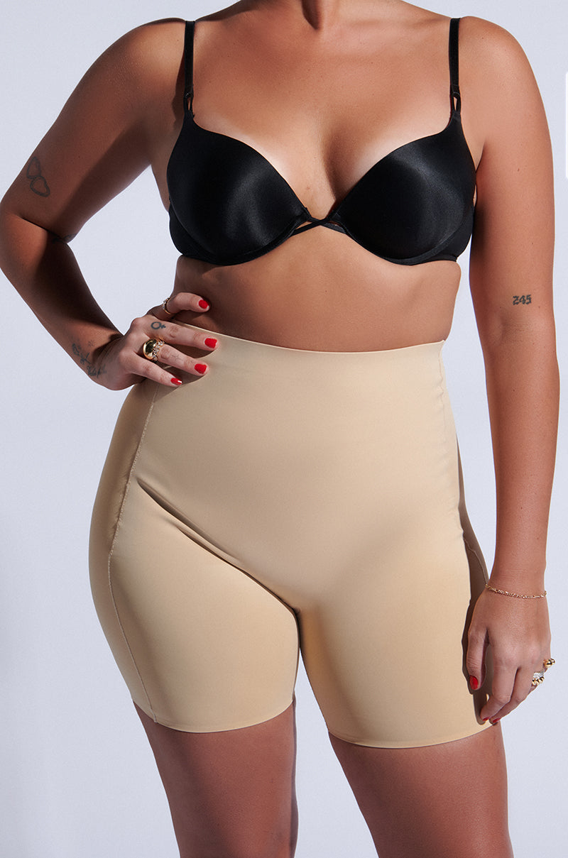 A person is wearing TITSY TUMMY TUCK SHORTS IN TAN with a black bra. Tattooed arms and red nail polish complement the look, set against a plain background. The image highlights their torso and upper legs. 5
